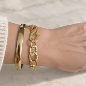 Gros plan sur un bracelet doré à maillons carrés entrelacés, aux angles arrondis et à la finition martelée, associé à un jonc lisse.