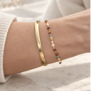 Gros plan sur un bracelet femme à petites pierres multicolores, perles blanches et détails dorés, associé à un jonc en acier inoxydable.