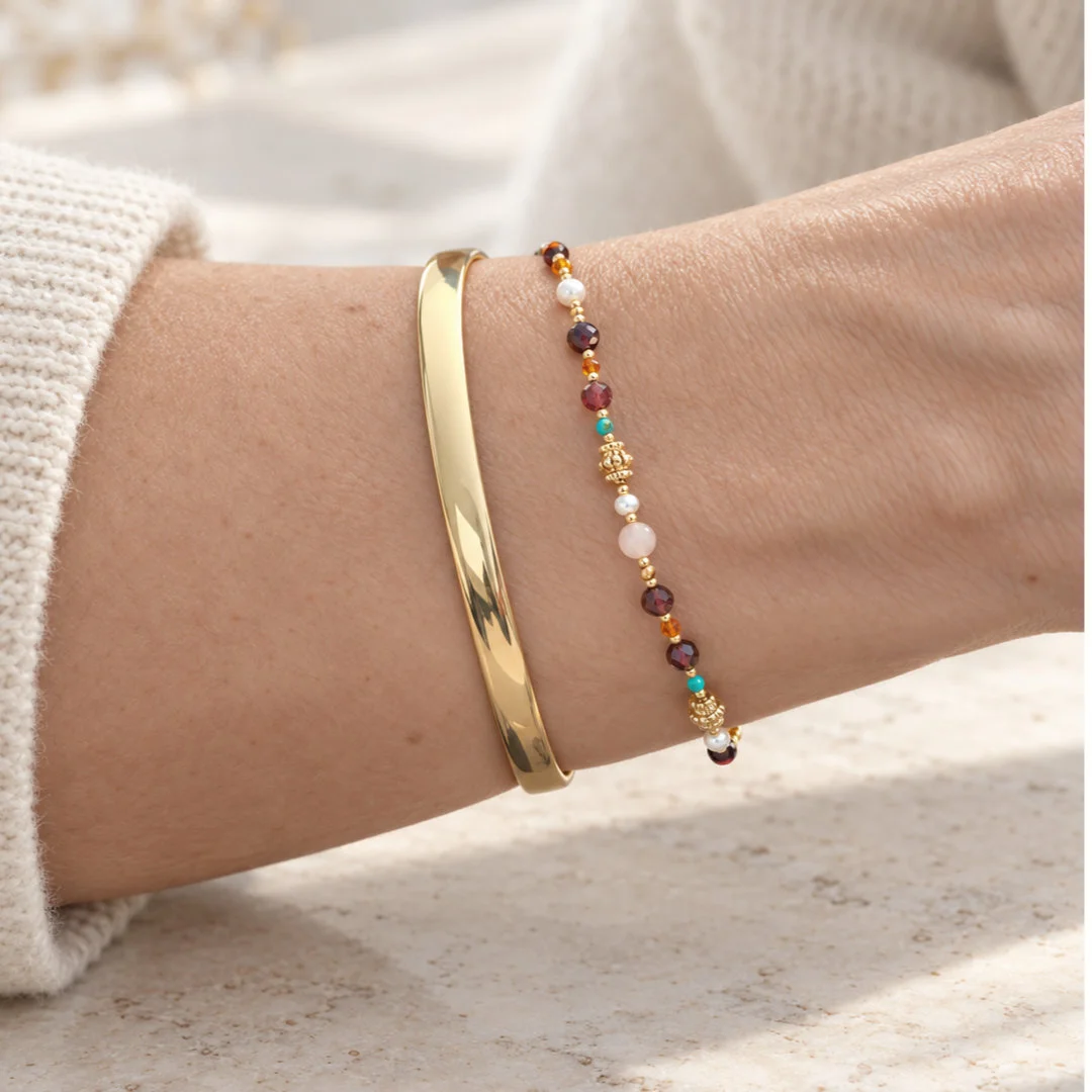 Gros plan sur un bracelet femme à petites pierres multicolores, perles blanches et détails dorés, associé à un jonc en acier inoxydable.