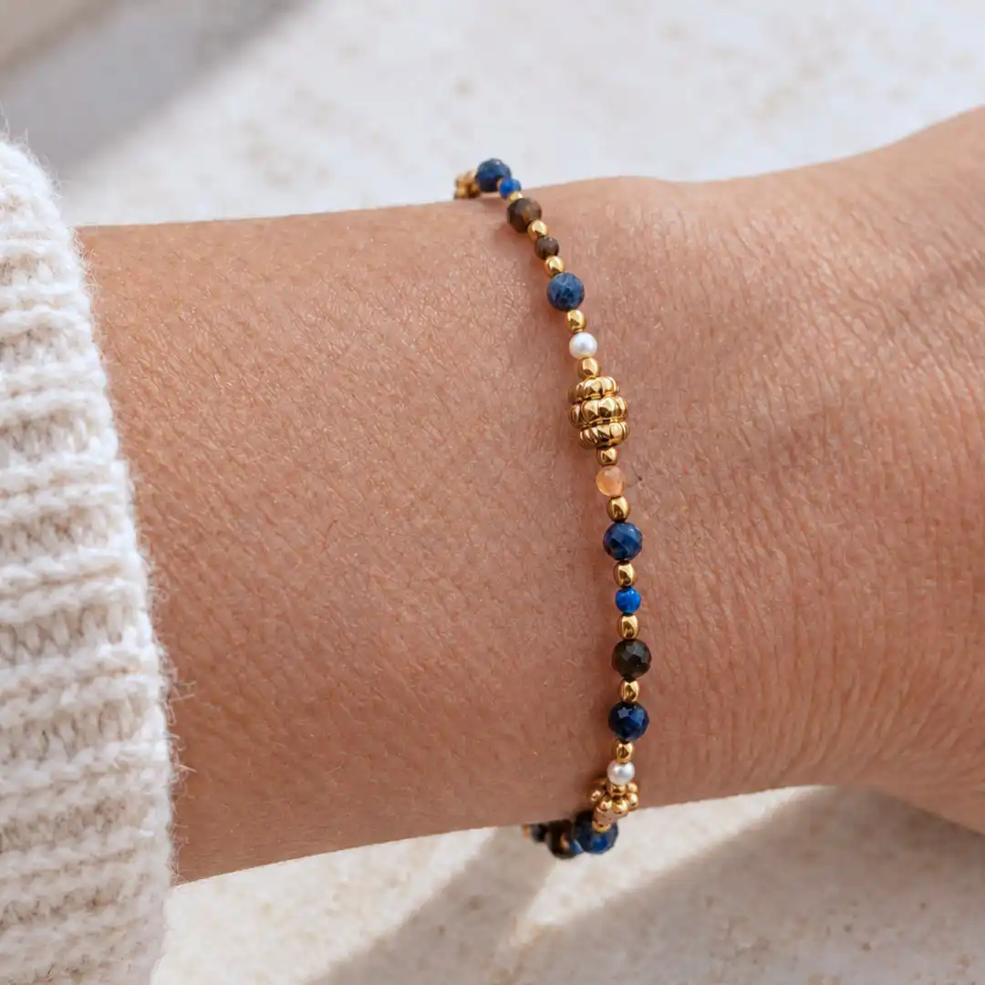 Bracelet femme à petites pierres bleues et brunes, perles blanches et détails dorés, avec perle centrale travaillée, au poignet.